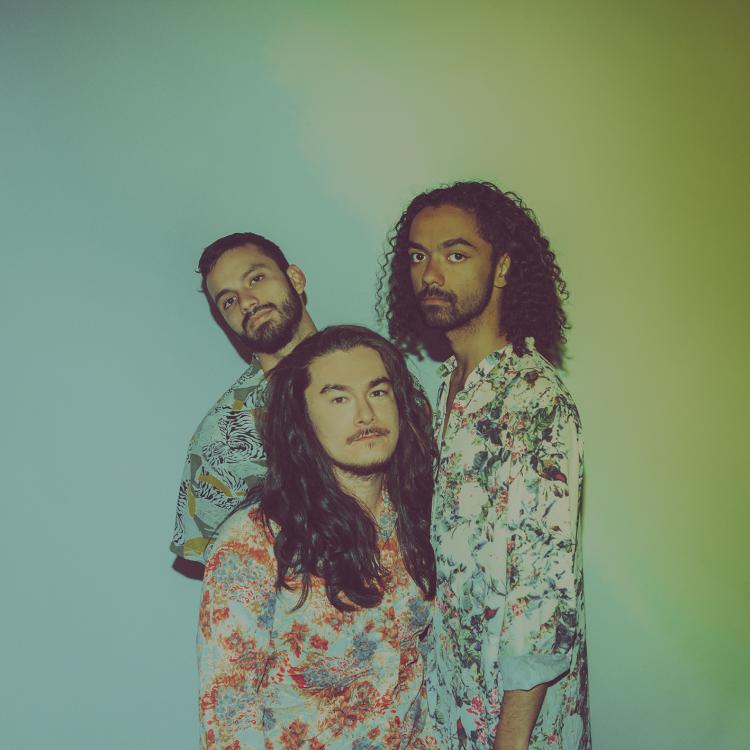 The three members of Izy wearing floral shirts standing in front of a blue and green gradient background