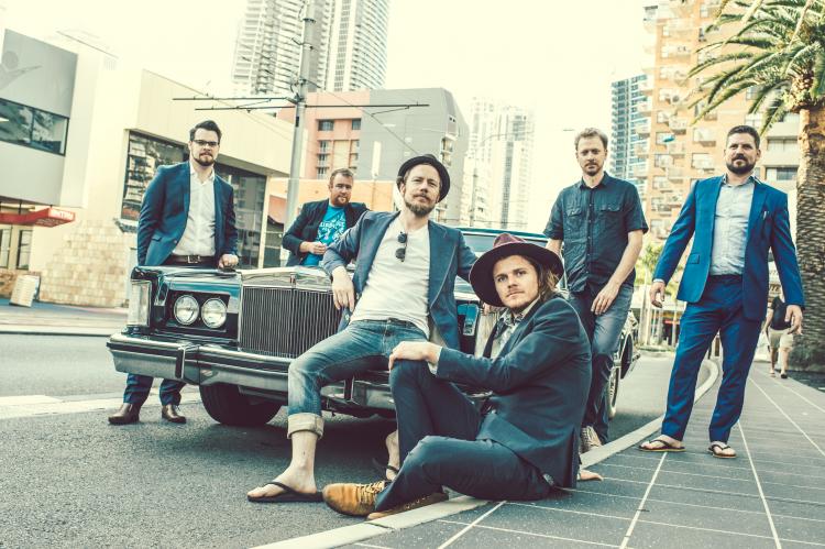 The members of Cheap Fakes sitting around an old school car wearing blue, alongside the sidewalk.
