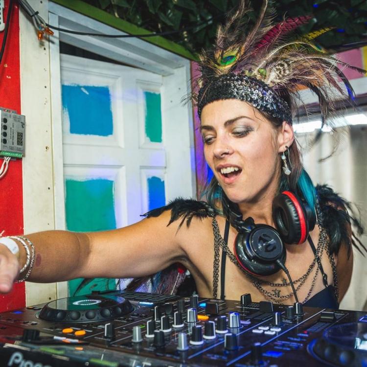 Mam Quilla DJing with a feathered headress, headphones around her neck and bead around her wrists. There is a pastel coloured door behind her.