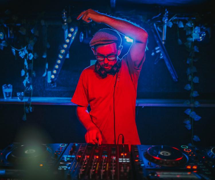 Gypsy Fingerz DJing in a button down shirt under red lights with his left hand in the air
